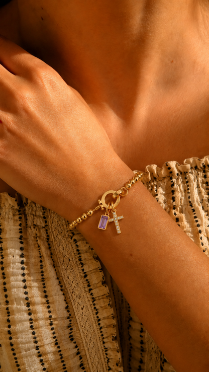 Pulseira dourada com contas e pingentes de cruz cravejada e pedra ametista retangular, em destaque no pulso sob luz quente.