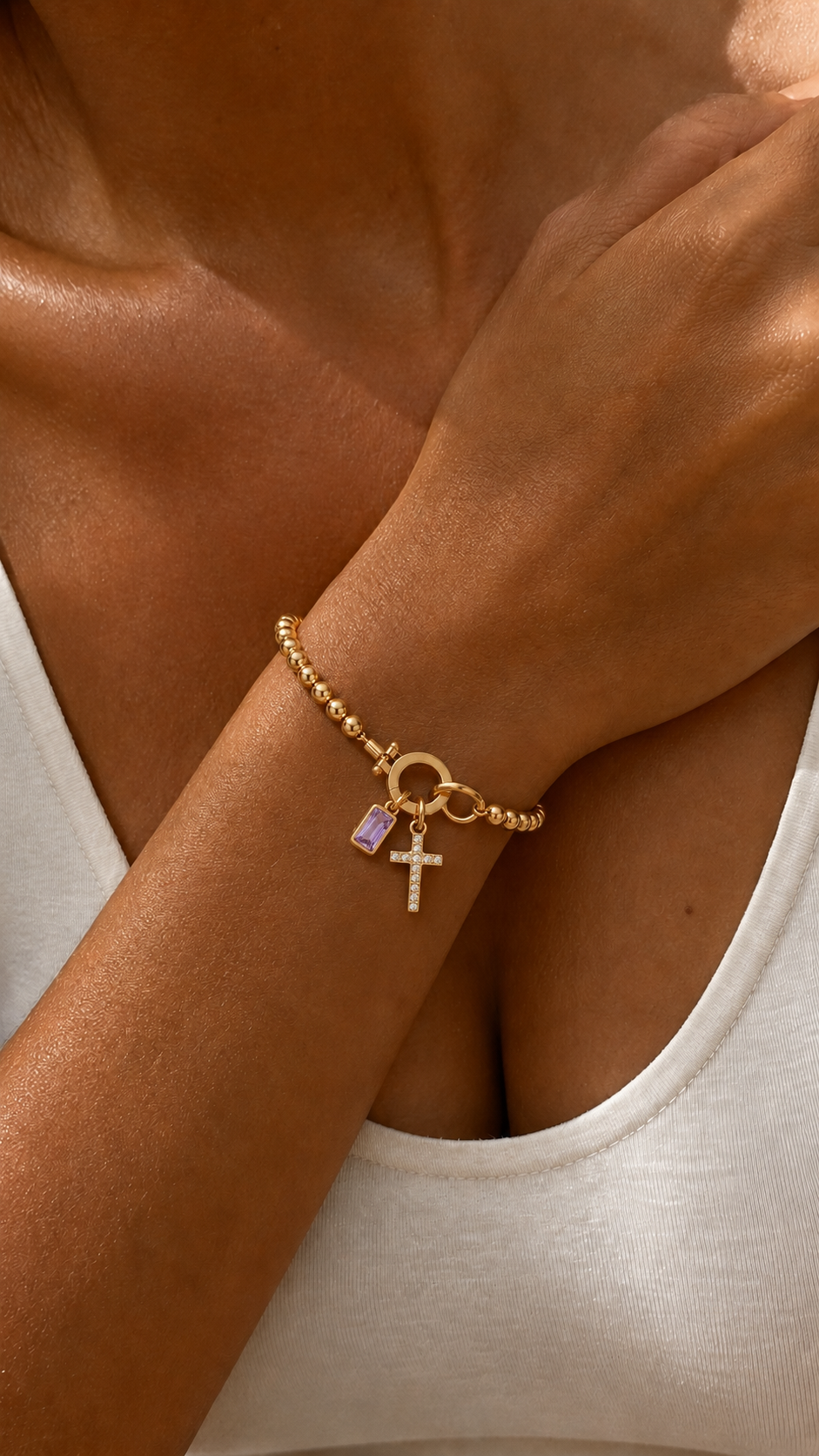 Pulseira dourada com contas e pingentes de cruz cravejada e pedra ametista retangular, em destaque no pulso sob luz quente.