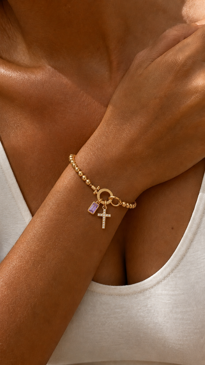Pulseira dourada com contas e pingentes de cruz cravejada e pedra ametista retangular, em destaque no pulso sob luz quente.