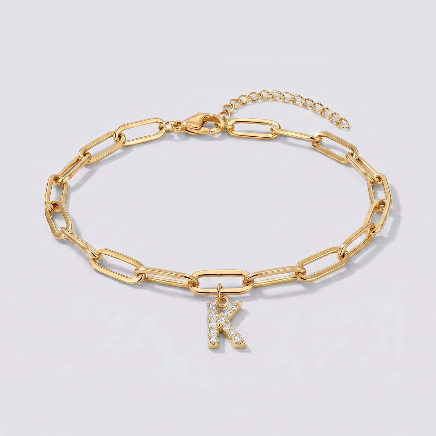 Pulseira dourada com letra inicial K, corrente paperclip e pendente de letra cravejado.