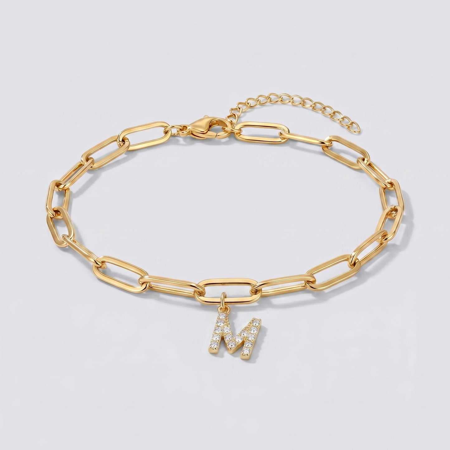 Pulseira dourada com letra inicial M, corrente paperclip e pendente de letra cravejado.