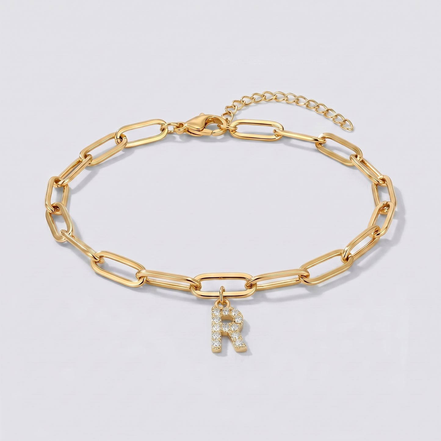 Pulseira dourada com letra inicial R, corrente paperclip e pendente de letra cravejado.