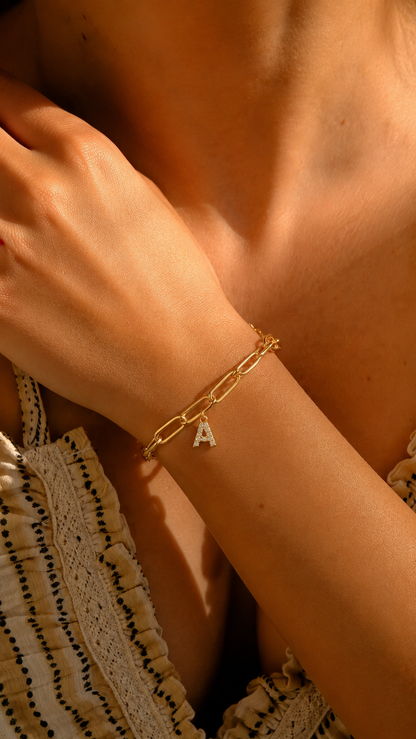 Pulseira dourada personalizada com inicial A, corrente paperclip e pendente de letra cravejado no pulso feminino.