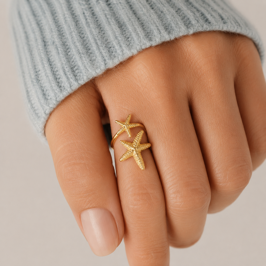 Anel ajustável dourado com duas estrelas-do-mar texturizadas usado no dedo, combinado com malha azul claro para um visual elegante, feminino e sofisticado.