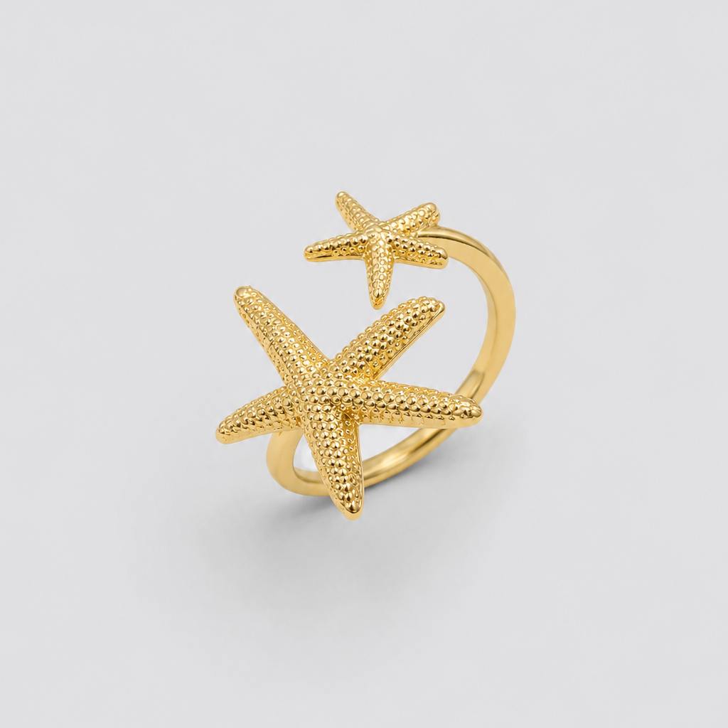 Anel ajustável dourado com duas estrelas-do-mar texturizadas em tamanhos diferentes, fotografado sobre fundo cinza claro para um visual elegante, minimalista e sofisticado.