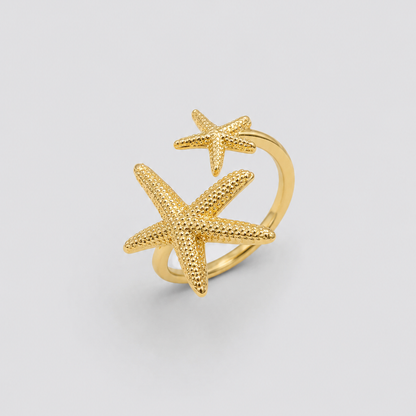 Anel ajustável dourado com duas estrelas-do-mar texturizadas em tamanhos diferentes, fotografado sobre fundo cinza claro para um visual elegante, minimalista e sofisticado.