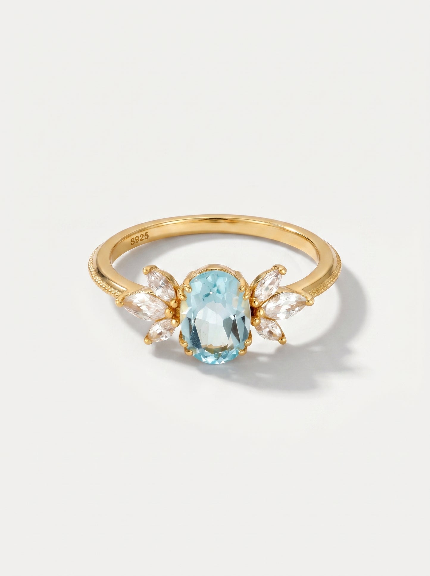Anel em ouro com uma pedra preciosa azul sobre um fundo cinzento claro.