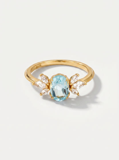 Anel em ouro com uma pedra preciosa azul sobre um fundo cinzento claro.