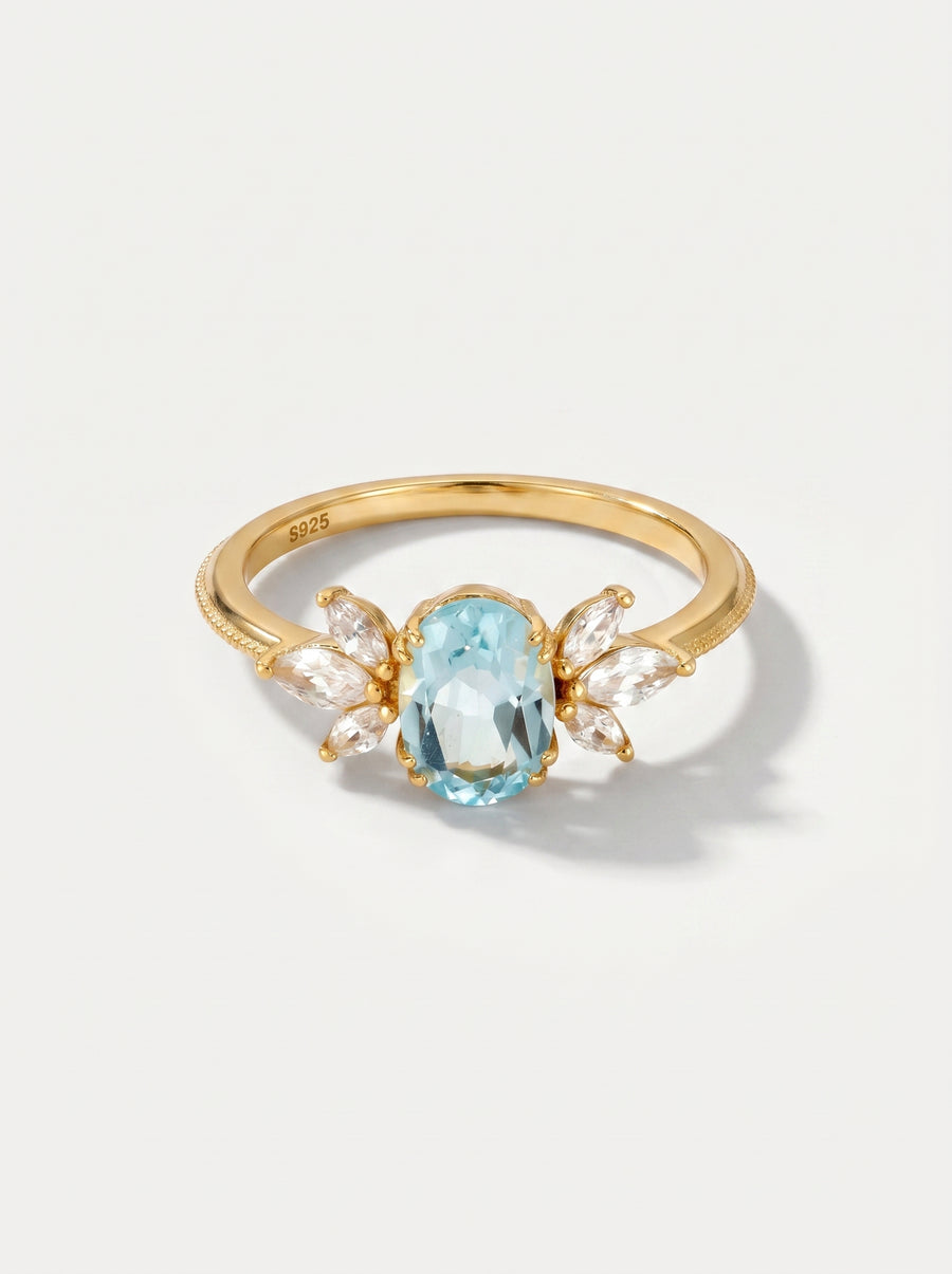 Anel em ouro com uma pedra preciosa azul sobre um fundo cinzento claro.