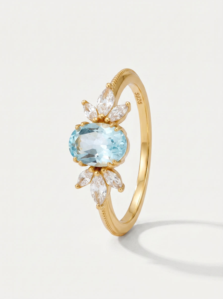 Anel em ouro com uma pedra preciosa azul e detalhes em diamante sobre um fundo cinzento claro.