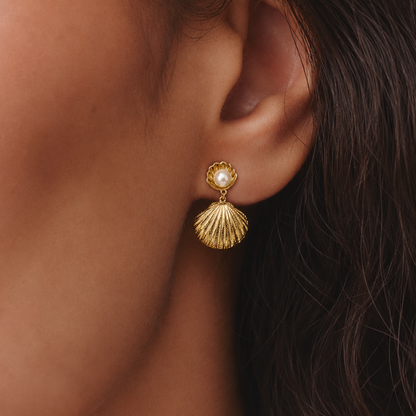 Brincos dourados com concha e pérola branca usados na orelha, para um visual elegante, feminino e sofisticado.