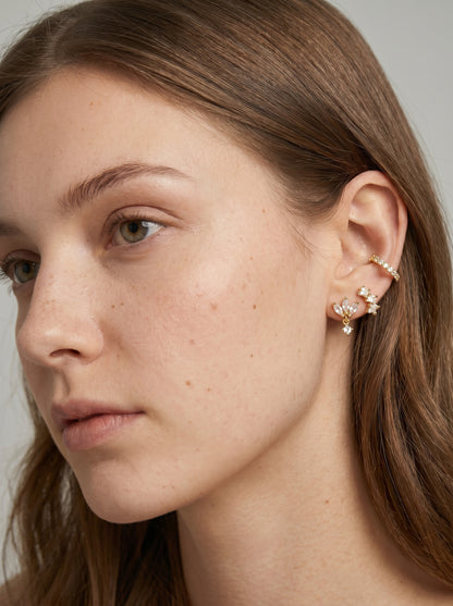 Brincos de pressão dourados e delicados brincos florais com detalhes em cristal transparente, exibidos numa modelo para um look elegante de joalharia.
