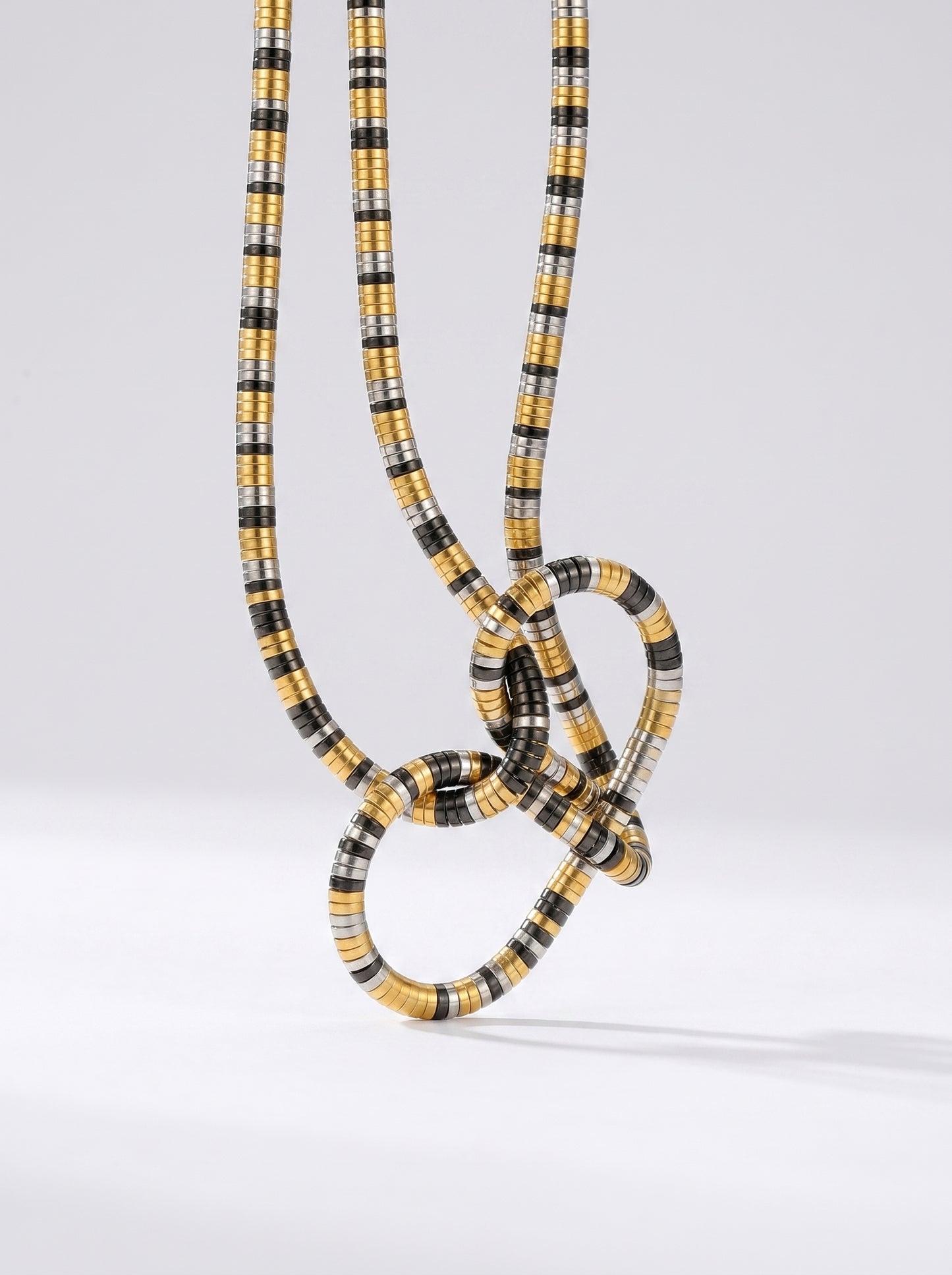 Colar bicolor em dourado, prateado e preto com pendente abstrato em forma de nó e acabamento texturizado, fotografado sobre fundo claro para um visual moderno, artístico e sofisticado.