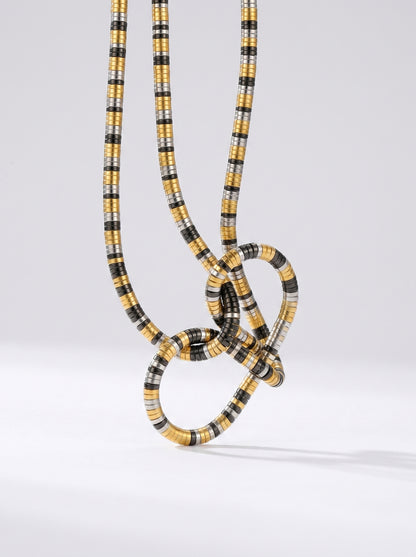 Colar bicolor em dourado, prateado e preto com pendente abstrato em forma de nó e acabamento texturizado, fotografado sobre fundo claro para um visual moderno, artístico e sofisticado.