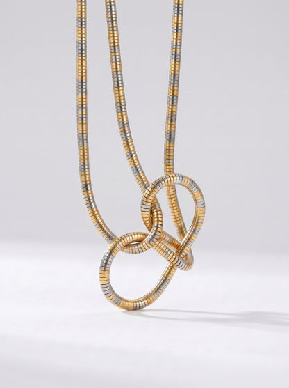 Colar bicolor em dourado e prateado com pendente abstrato em forma de nó e acabamento texturizado, fotografado sobre fundo claro para um visual moderno, artístico e sofisticado.