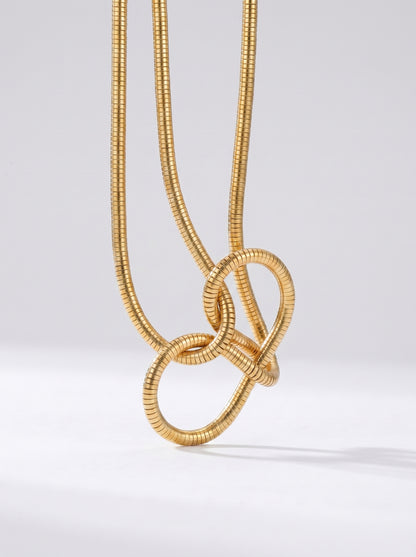 Colar dourado moderno com pendente abstrato em forma de nó e acabamento texturizado, fotografado sobre fundo claro para um visual elegante, artístico e sofisticado.