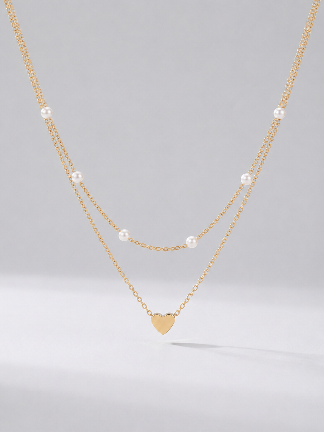 Colar dourado delicado em duas camadas com pérolas brancas e pendente em forma de coração, fotografado sobre fundo claro para um visual elegante, romântico e sofisticado.