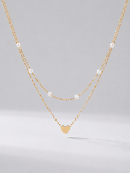 Colar dourado delicado em duas camadas com pérolas brancas e pendente em forma de coração, fotografado sobre fundo claro para um visual elegante, romântico e sofisticado.