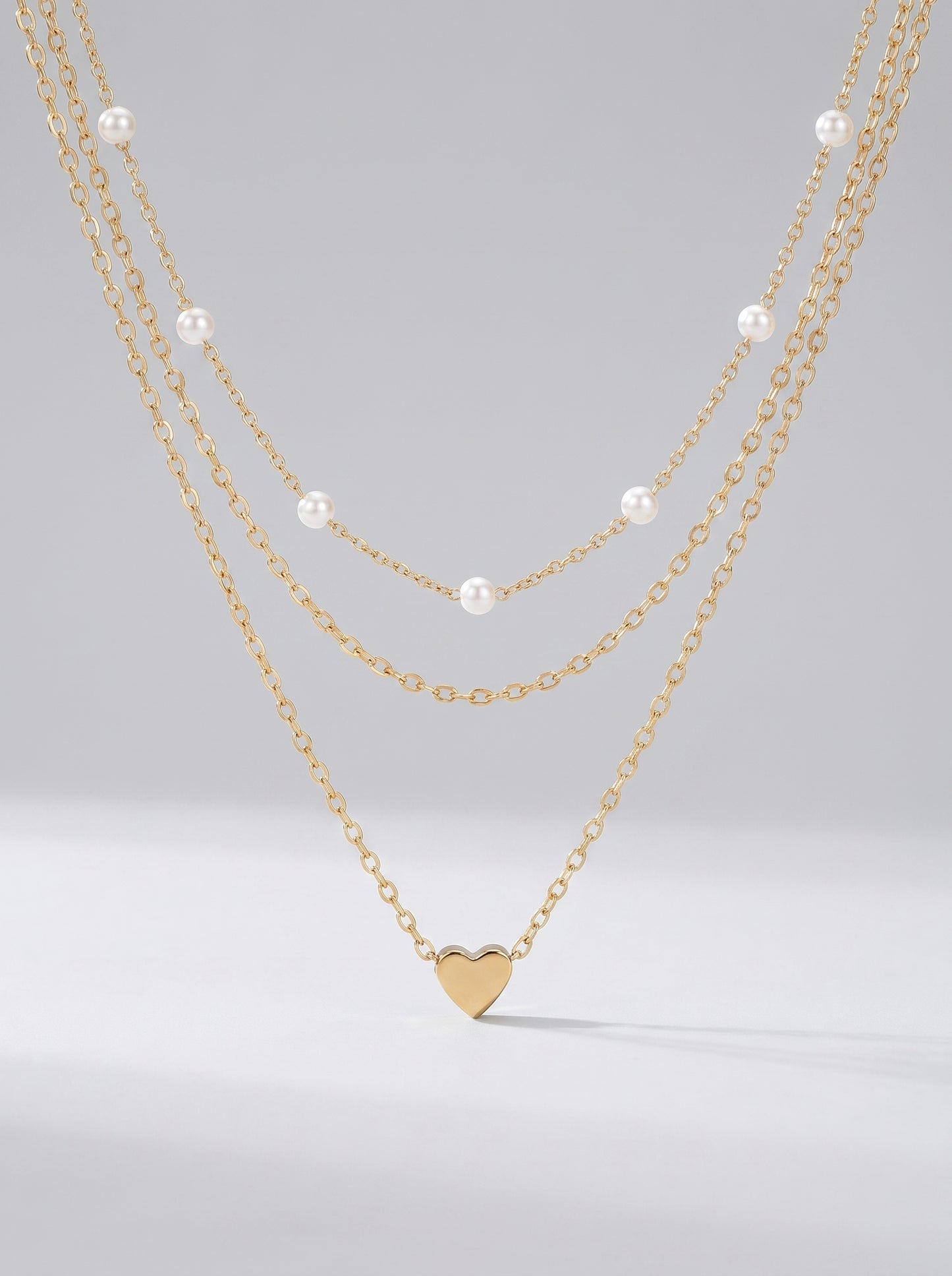 Colar dourado delicado em três camadas com pérolas brancas e pendente em forma de coração, fotografado sobre fundo claro para um visual elegante, romântico e sofisticado.