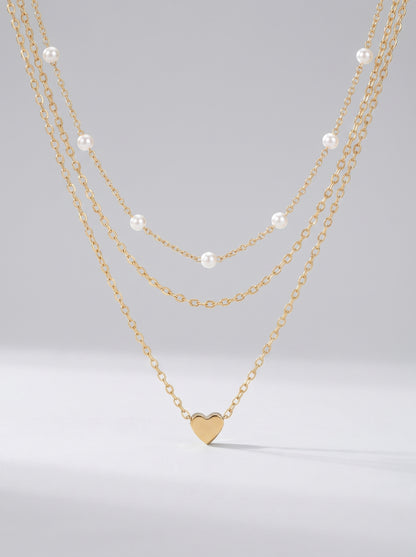 Colar dourado delicado em três camadas com pérolas brancas e pendente em forma de coração, fotografado sobre fundo claro para um visual elegante, romântico e sofisticado.