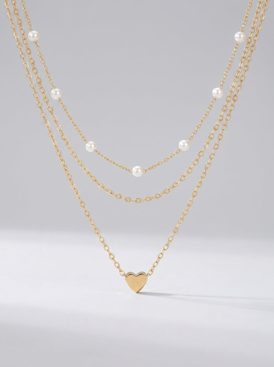 Colar dourado delicado em três camadas com pérolas brancas e pendente em forma de coração, fotografado sobre fundo claro para um visual elegante, romântico e sofisticado.