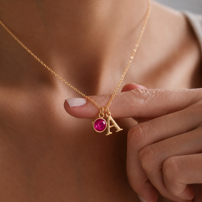 Close-up de colar dourado com inicial A e pedra de nascimento rosa, segurado à mão para destacar os detalhes personalizados.
