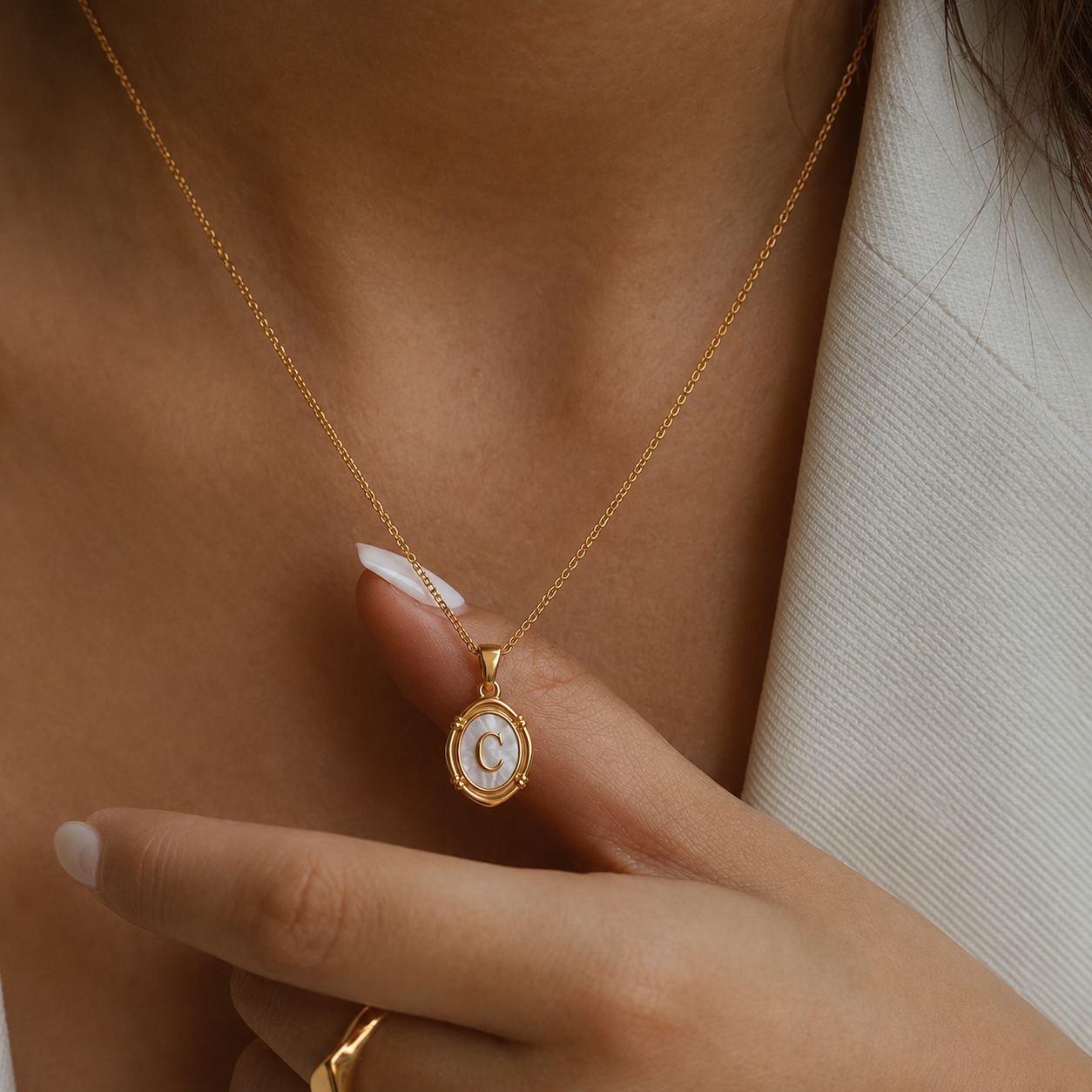 Close-up de colar oval dourado com inicial C em madrepérola, usado ao pescoço e destacado pela mão.