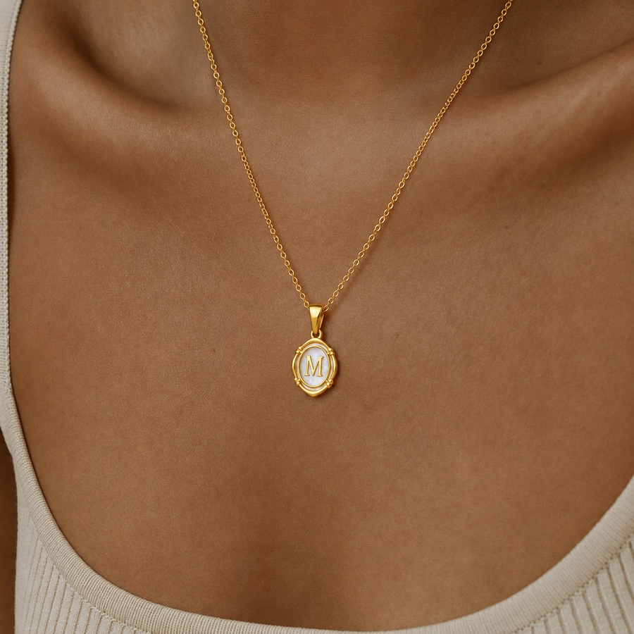 Mulher a usar colar oval dourado personalizado com inicial M em madrepérola sobre o pescoço.