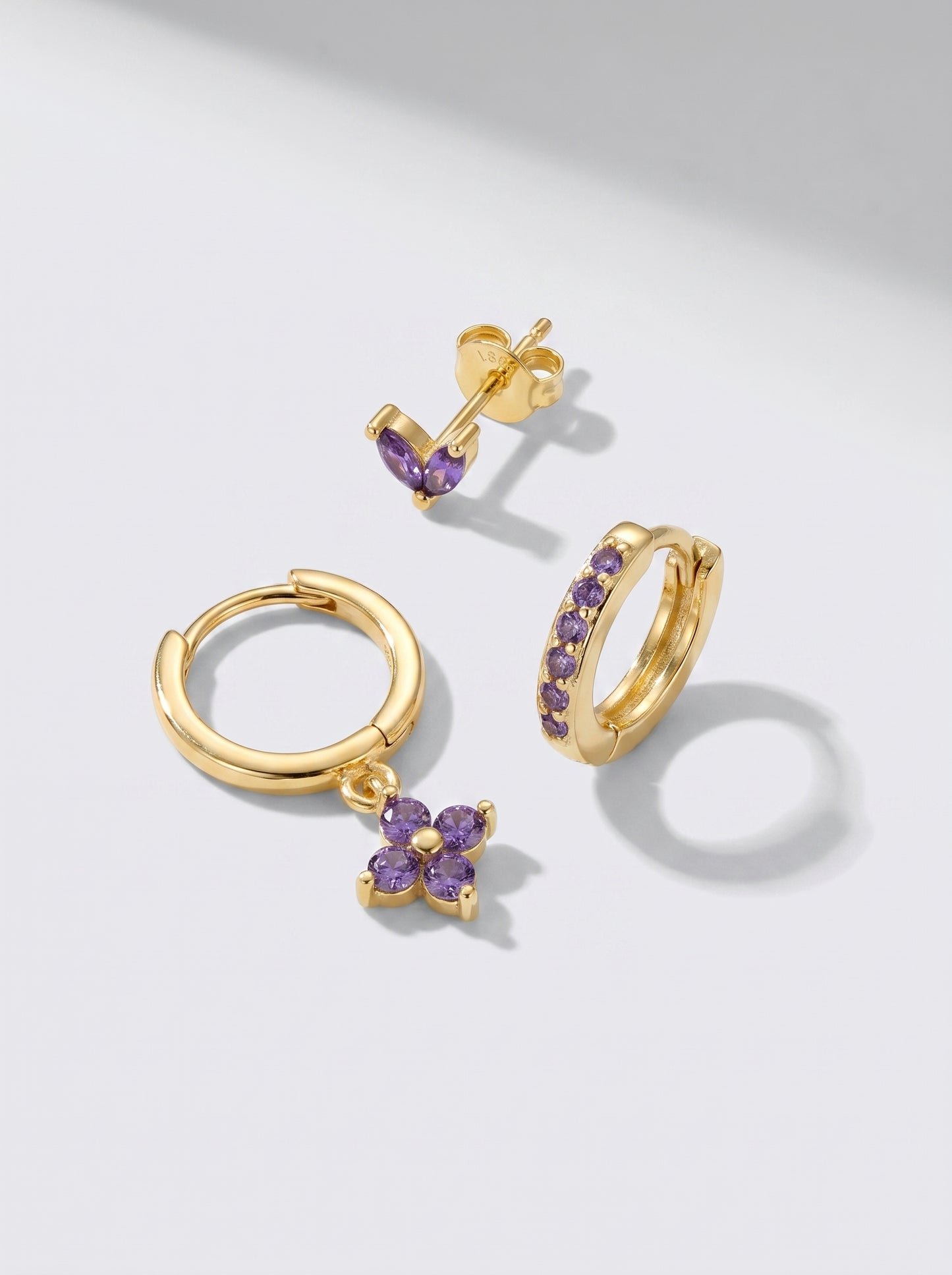 Gold hoop earrings and stud earrings with purple crystal accents, photographed on a minimalist light grey background.