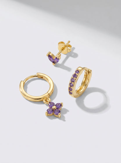 Gold hoop earrings and stud earrings with purple crystal accents, photographed on a minimalist light grey background.