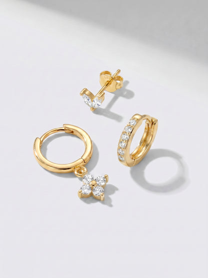 Gold hoop earrings and stud earrings with white crystal accents, photographed on a minimalist light grey background.