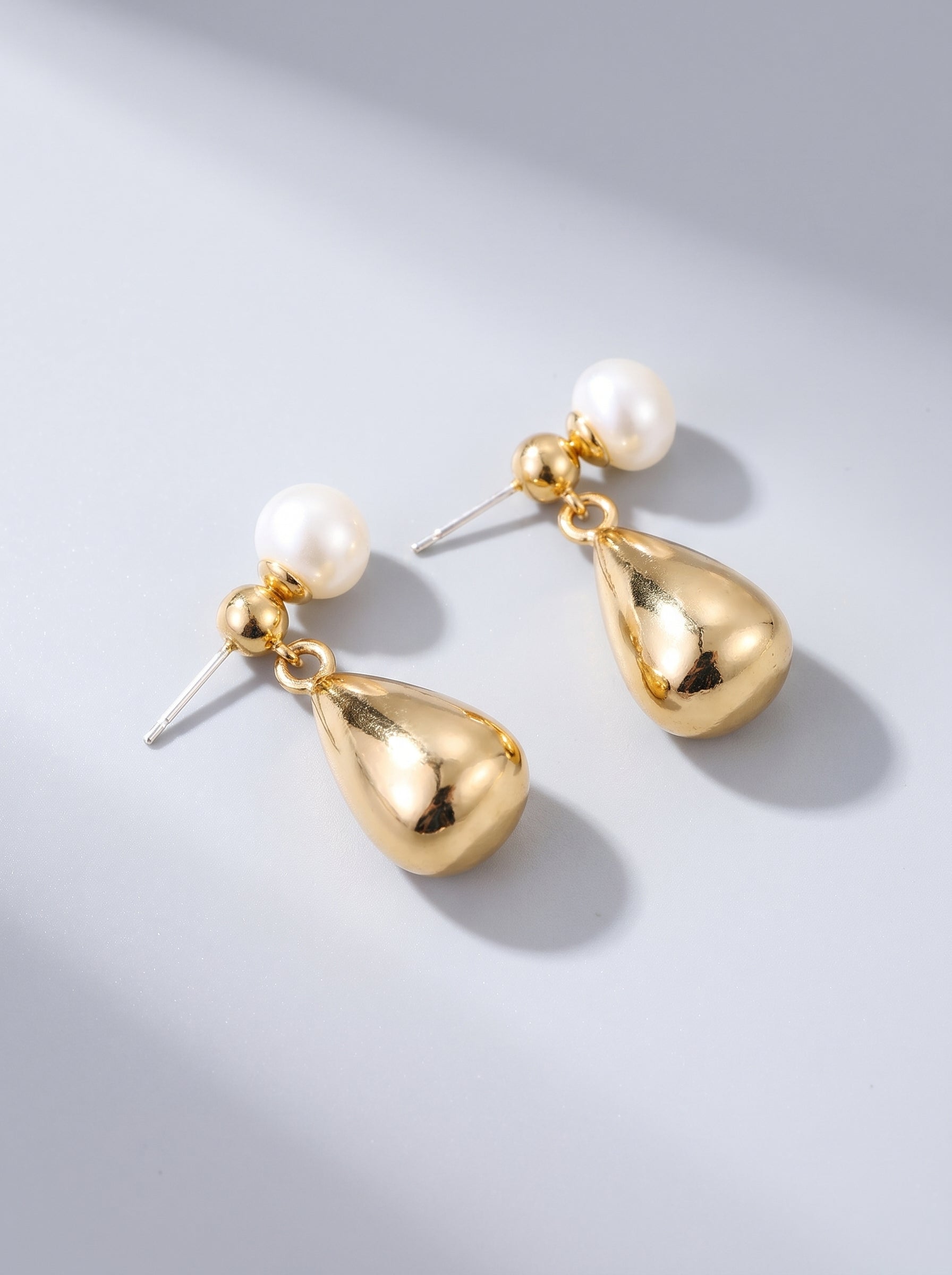 Brincos dourados pendentes com pérola branca e gota metálica, fotografados sobre fundo cinzento claro para um visual elegante e sofisticado.