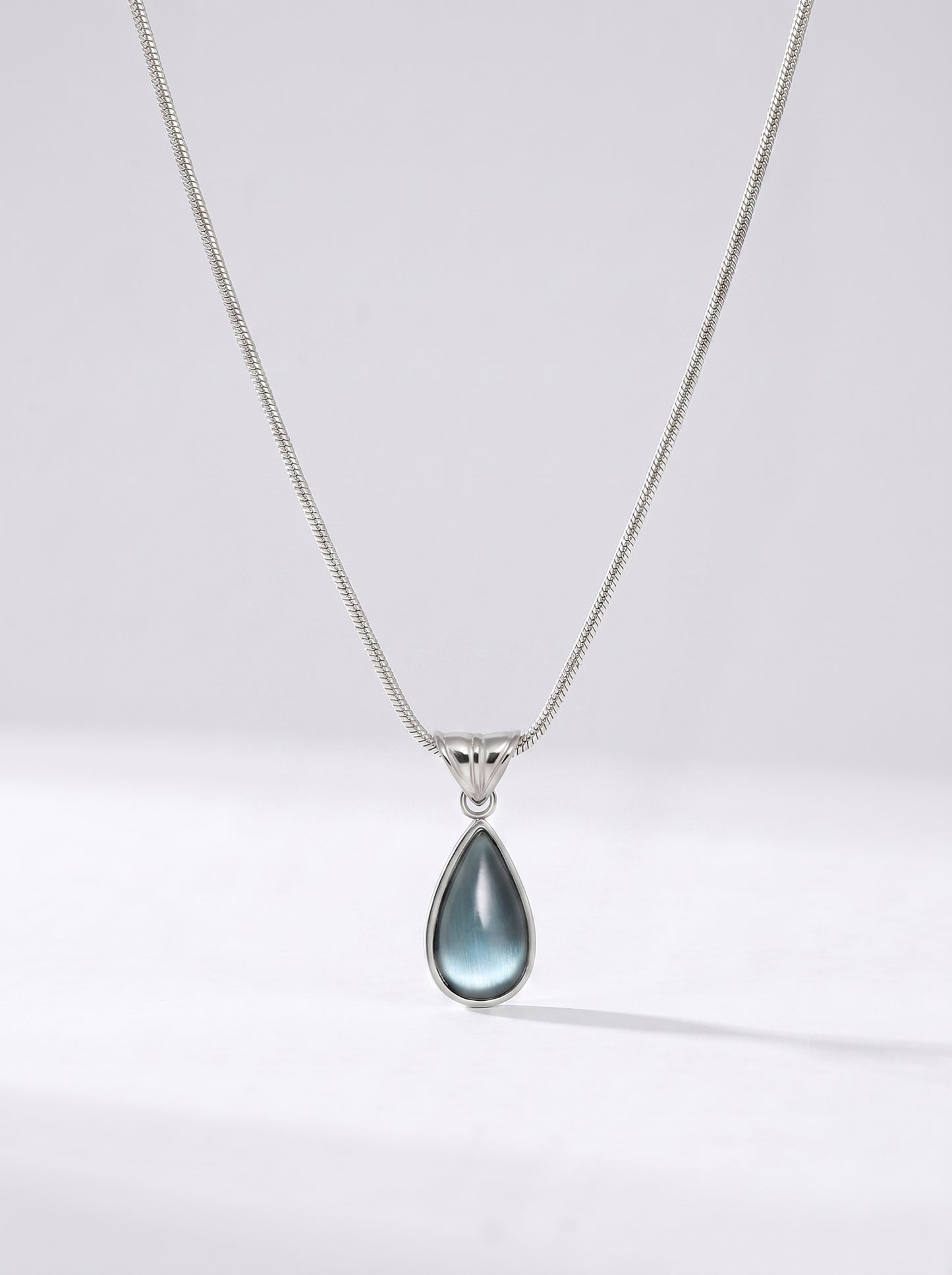 Colar prateado delicado com pendente azul em forma de gota e acabamento elegante, fotografado sobre fundo claro para um visual sofisticado, moderno e delicado.