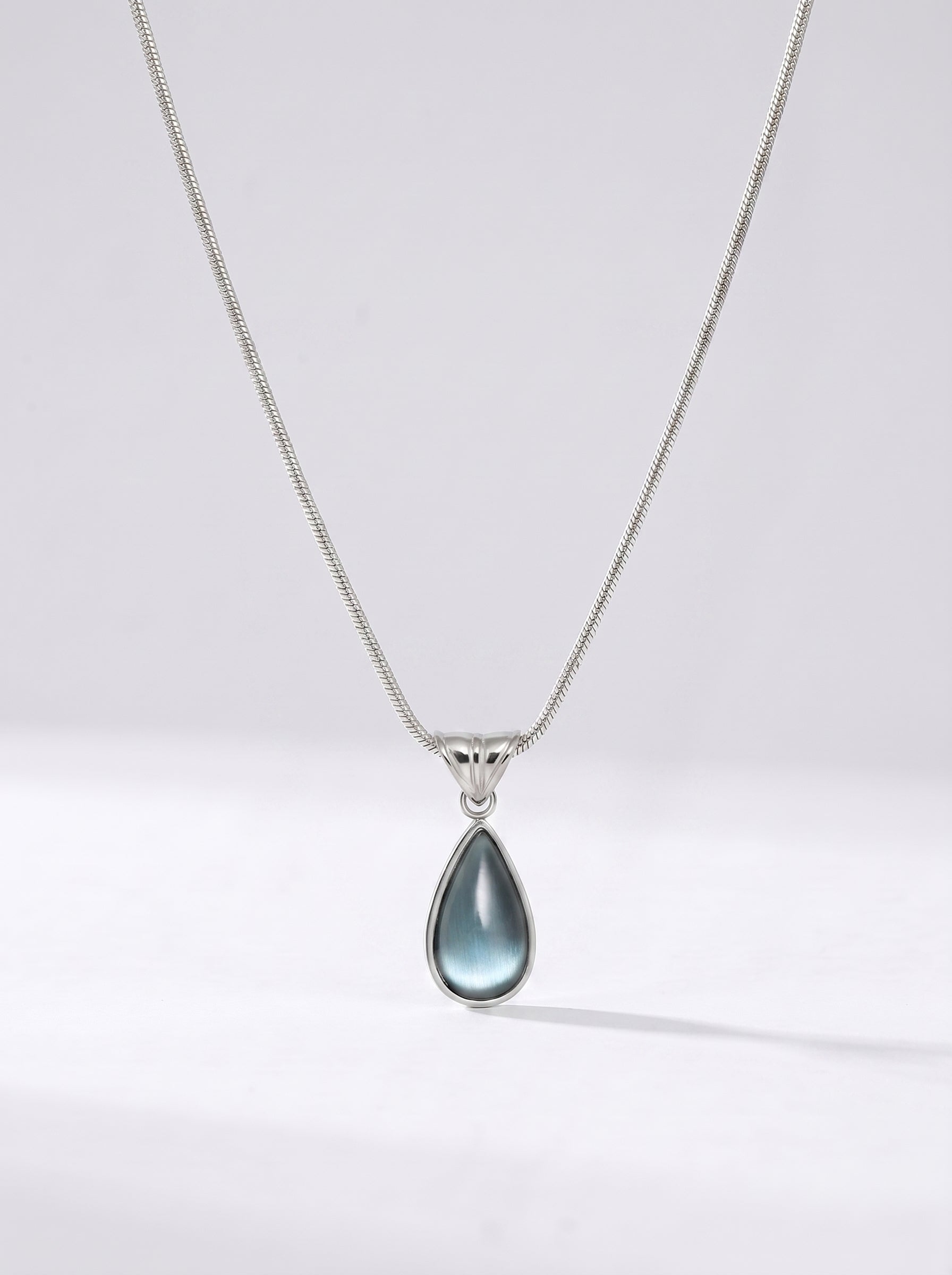 Colar prateado delicado com pendente azul em forma de gota e acabamento elegante, fotografado sobre fundo claro para um visual sofisticado, moderno e delicado.