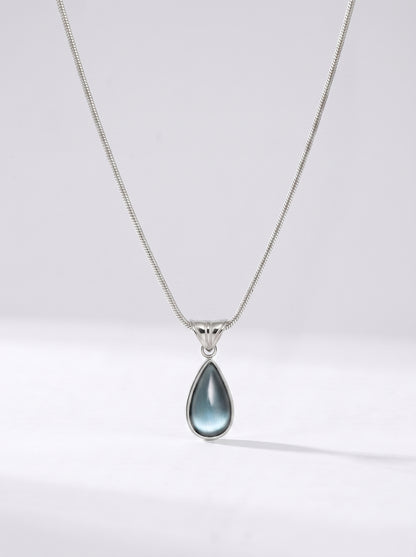 Colar prateado delicado com pendente azul em forma de gota e acabamento elegante, fotografado sobre fundo claro para um visual sofisticado, moderno e delicado.