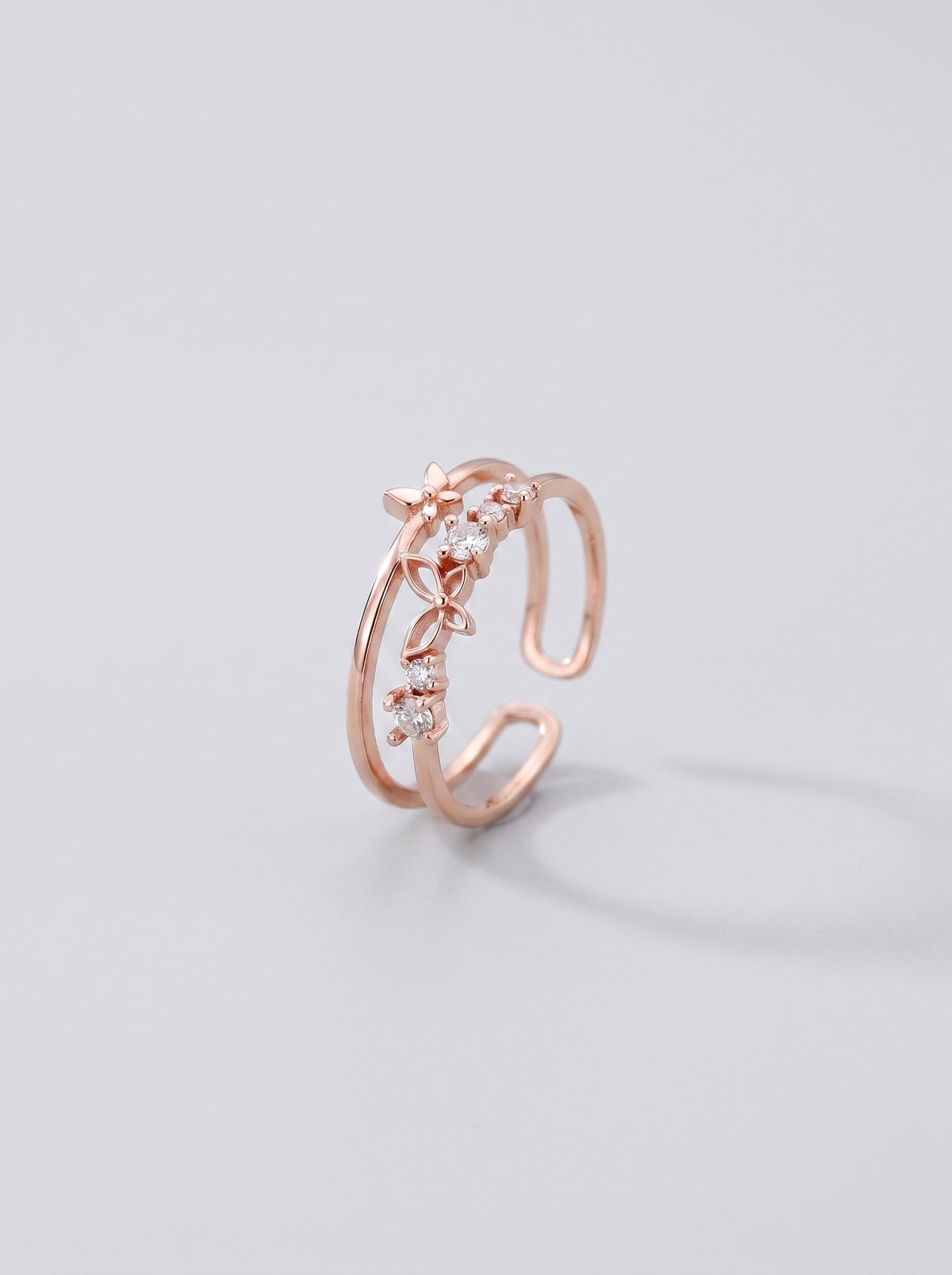 Anel aberto em ouro rosa com desenho floral sobre fundo cinza claro.
