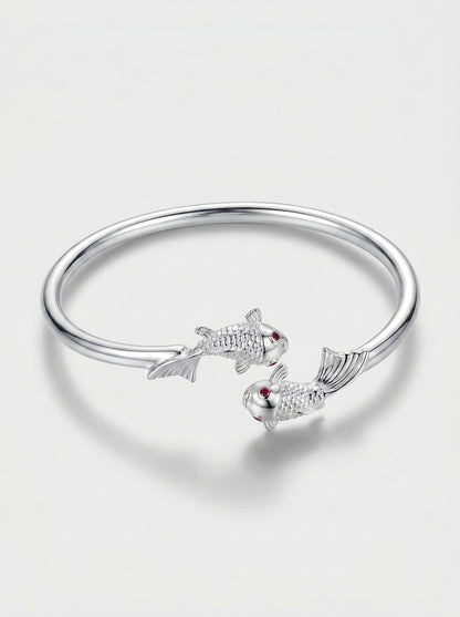 Pulseira prateada rígida com design aberto e detalhes delicados de peixes, fotografada sobre fundo claro para um visual elegante e minimalista.