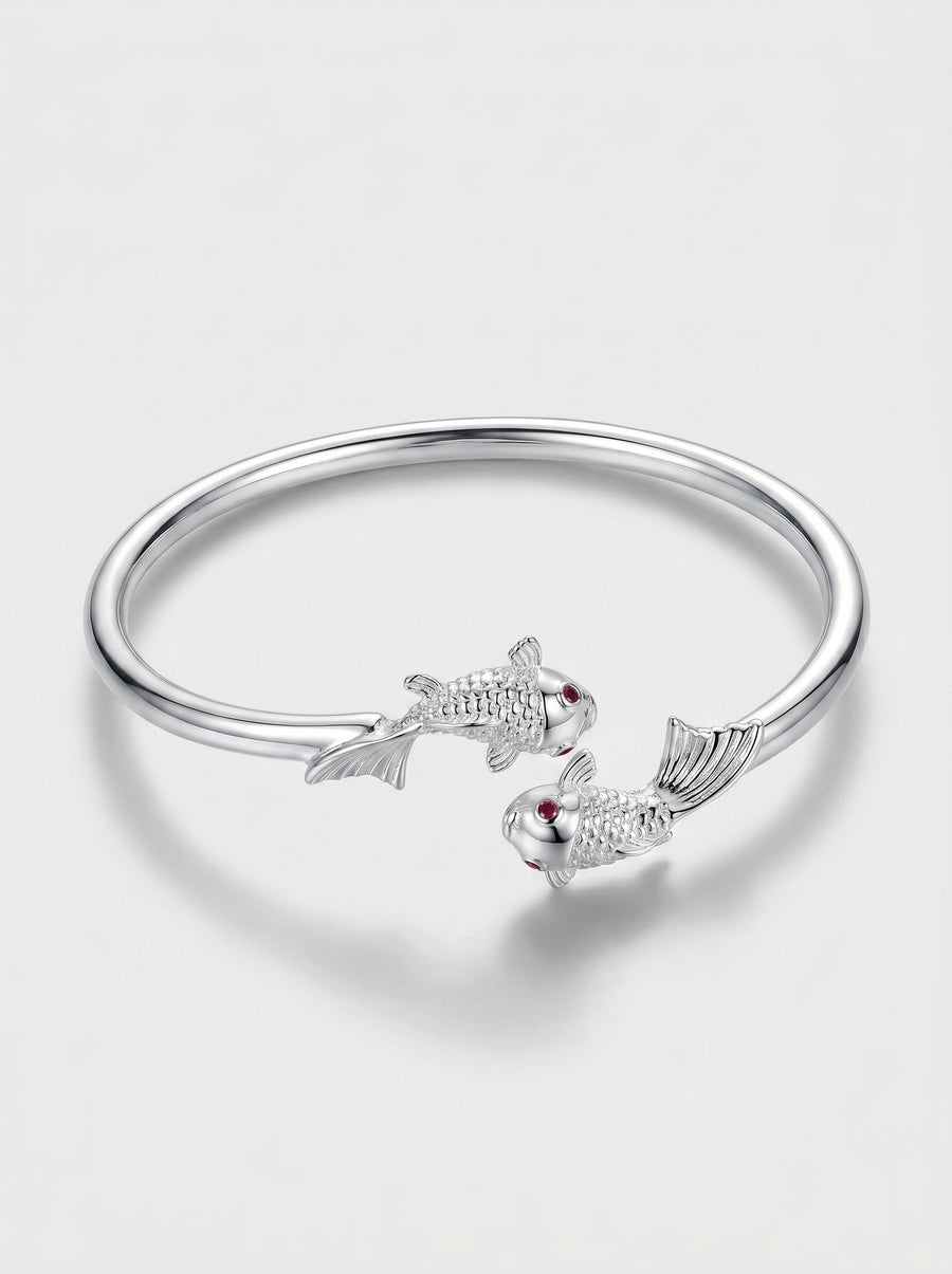 Pulseira prateada rígida com design aberto e detalhes delicados de peixes, fotografada sobre fundo claro para um visual elegante e minimalista.