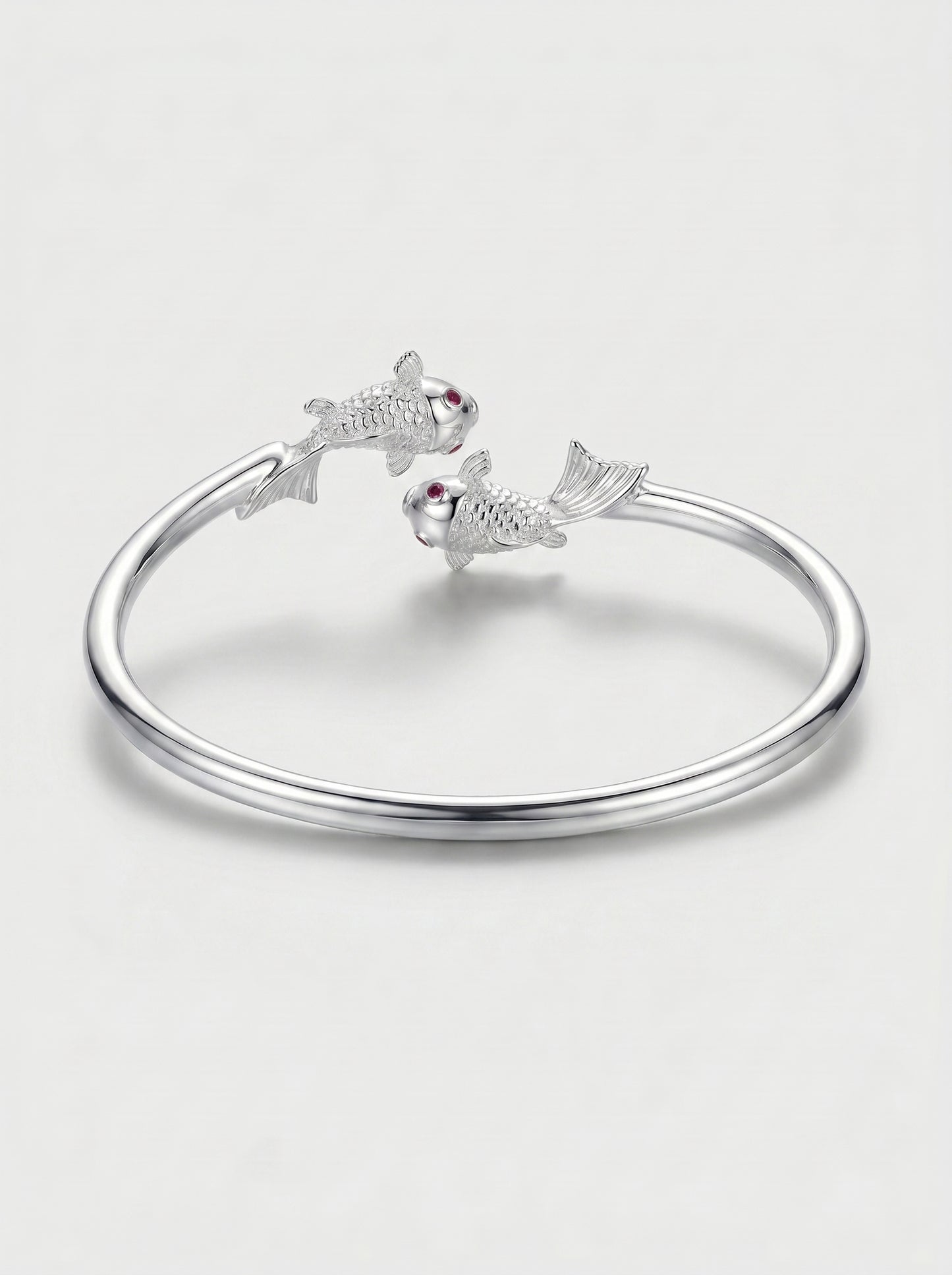 Pulseira prateada rígida com design aberto e detalhes de dois peixes nas extremidades, fotografada sobre fundo claro para um visual elegante e delicado.