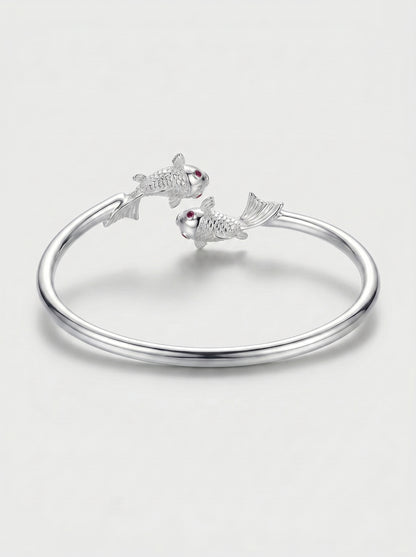 Pulseira prateada rígida com design aberto e detalhes de dois peixes nas extremidades, fotografada sobre fundo claro para um visual elegante e delicado.