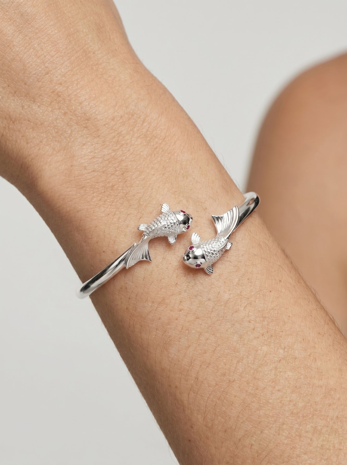 Pulseira prateada rígida com design aberto e detalhes de peixes nas extremidades, usada no pulso para um visual elegante e delicado.