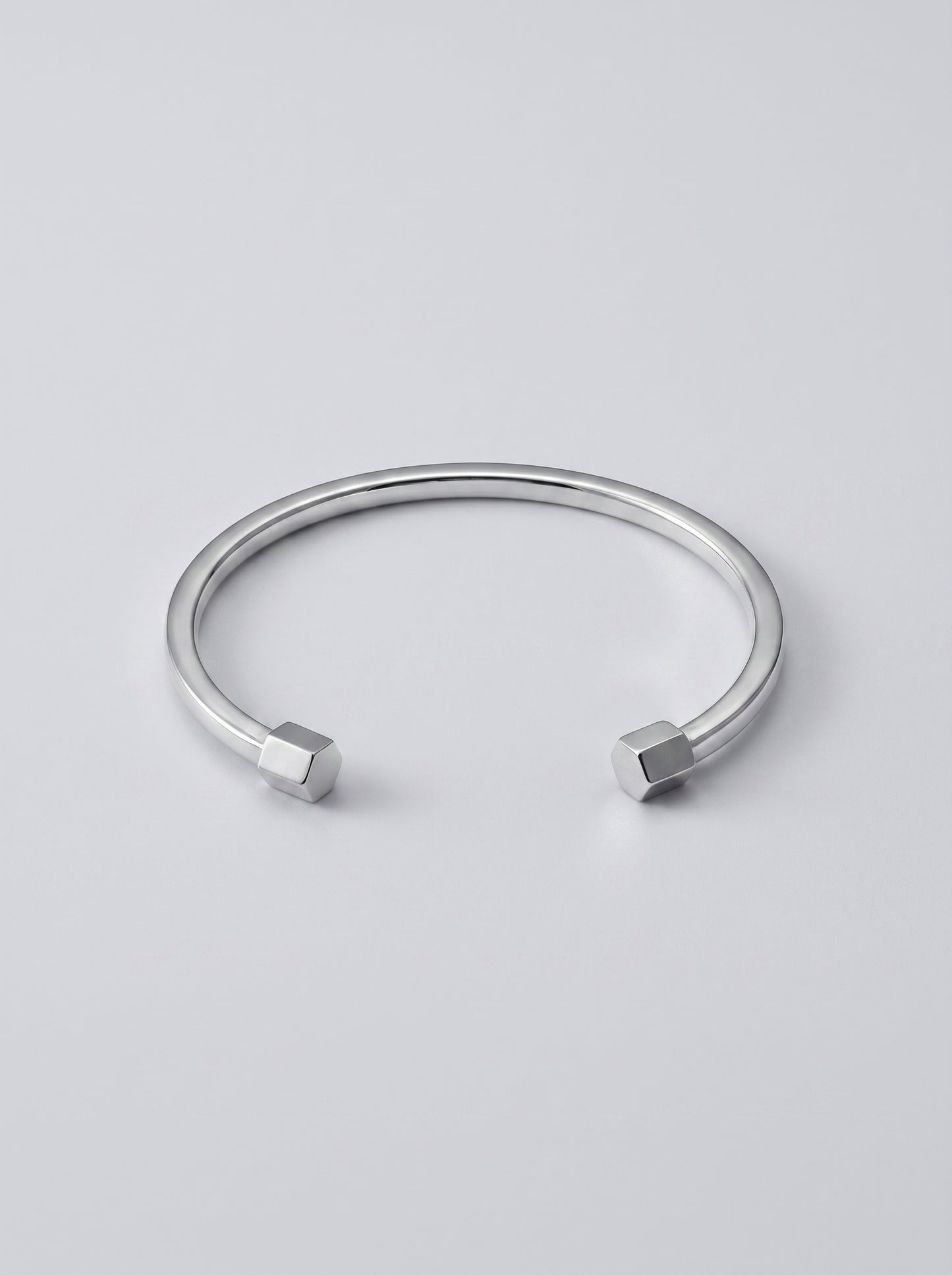 Pulseira prateada rígida com design aberto e detalhes geométricos nas extremidades, fotografada sobre fundo cinzento claro para um visual minimalista e moderno.