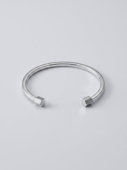 Pulseira prateada rígida com design aberto e detalhes geométricos nas extremidades, fotografada sobre fundo cinzento claro para um visual minimalista e moderno.