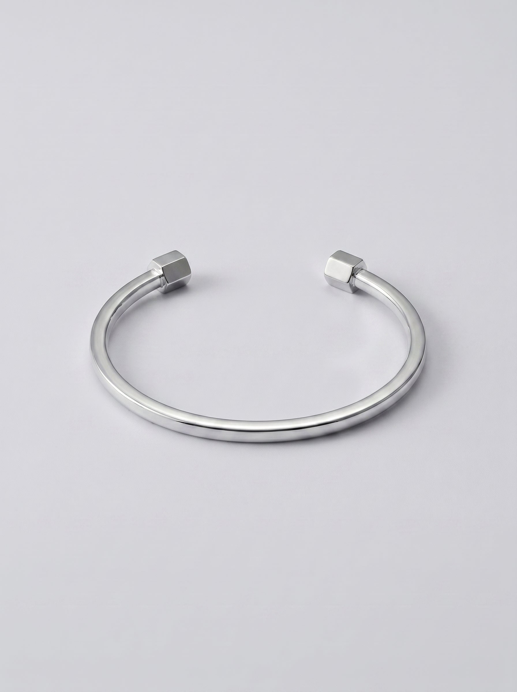 Pulseira prateada aberta com extremidades em forma de hexagono sobre fundo cinzento claro.