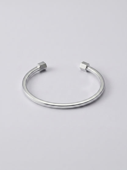 Pulseira prateada aberta com extremidades em forma de hexagono sobre fundo cinzento claro.