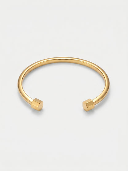 Pulseira dourada rígida com design aberto, acabamento polido e extremidades hexagonais, fotografada sobre fundo claro para um visual minimalista e moderno.