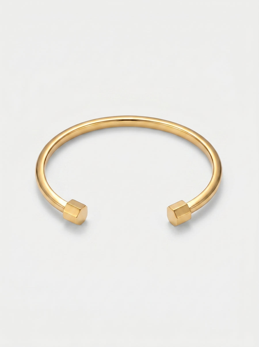 Pulseira dourada rígida com design aberto, acabamento polido e extremidades hexagonais, fotografada sobre fundo claro para um visual minimalista e moderno.