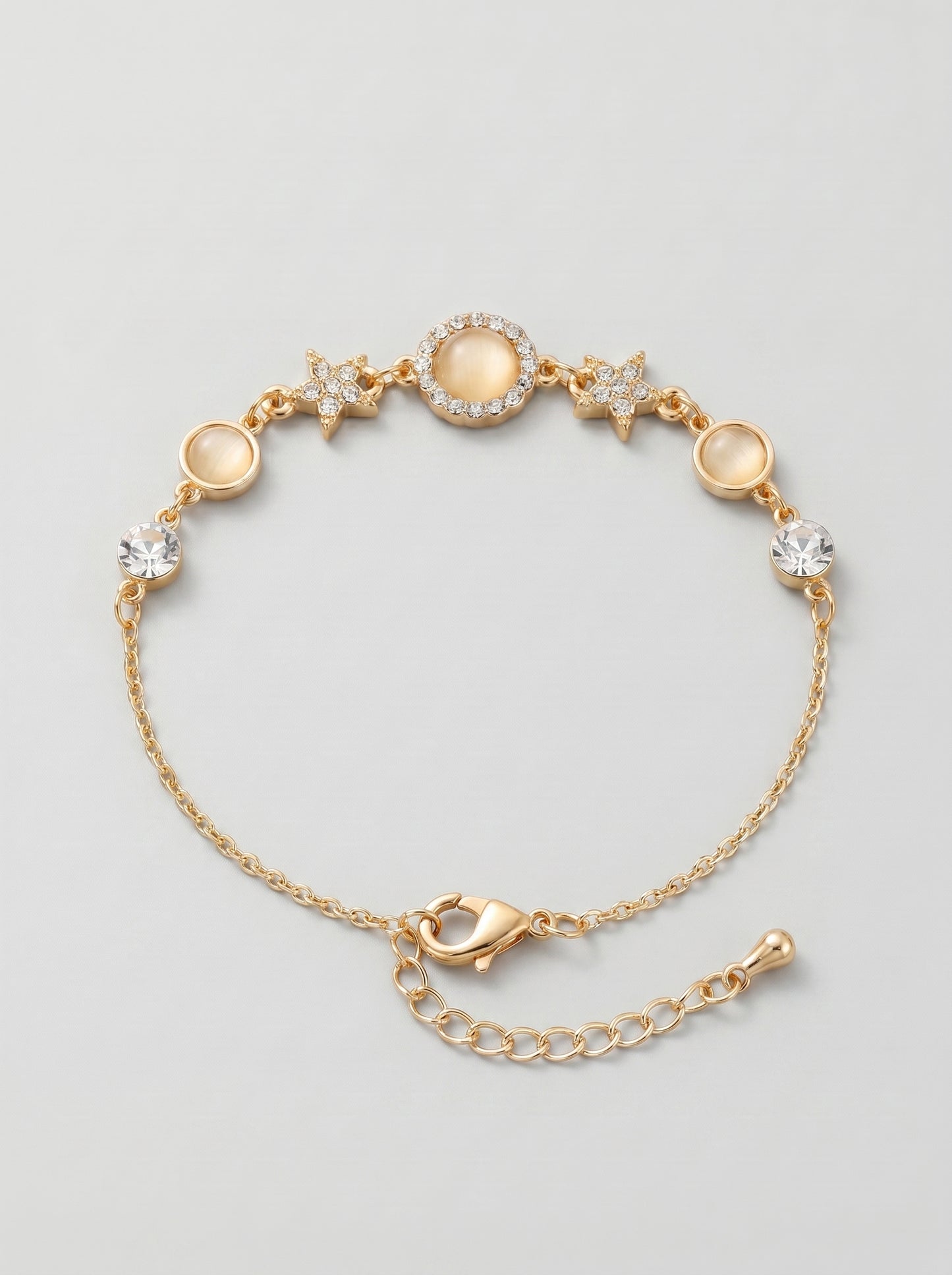 Pulseira dourada delicada com cristais brilhantes, detalhes em estrela e pendente circular central, fotografada sobre fundo cinzento claro para um visual elegante, feminino e sofisticado.