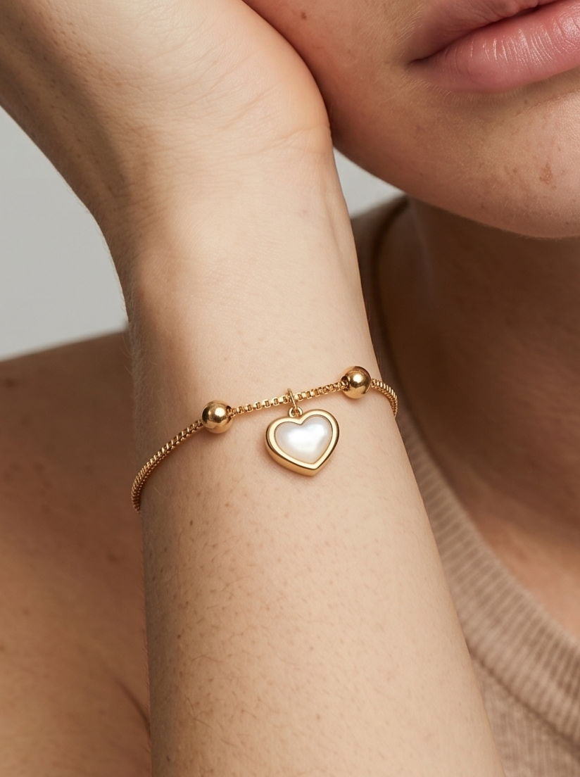 Pulseira dourada delicada com pendente em forma de coração branco e acabamento brilhante, usada no pulso para um visual elegante, feminino e romântico.