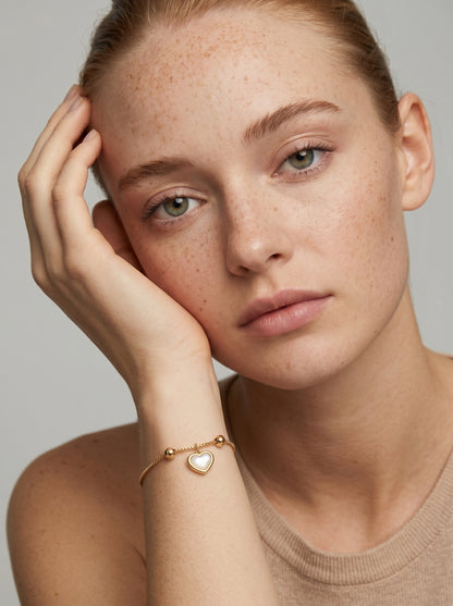 Pulseira dourada delicada com pendente em forma de coração branco e acabamento brilhante, usada por uma modelo para um visual elegante, feminino e romântico.